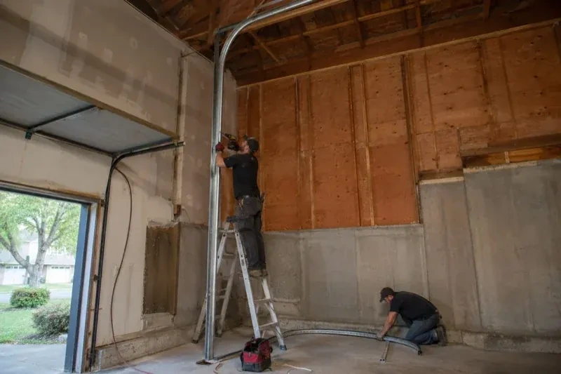 One person on a stepladder working beside a tall curved garage-door track while another kneels beside loose track sections in an unfinished garage. — High-Lift Garage Door Conversion, Burlington
